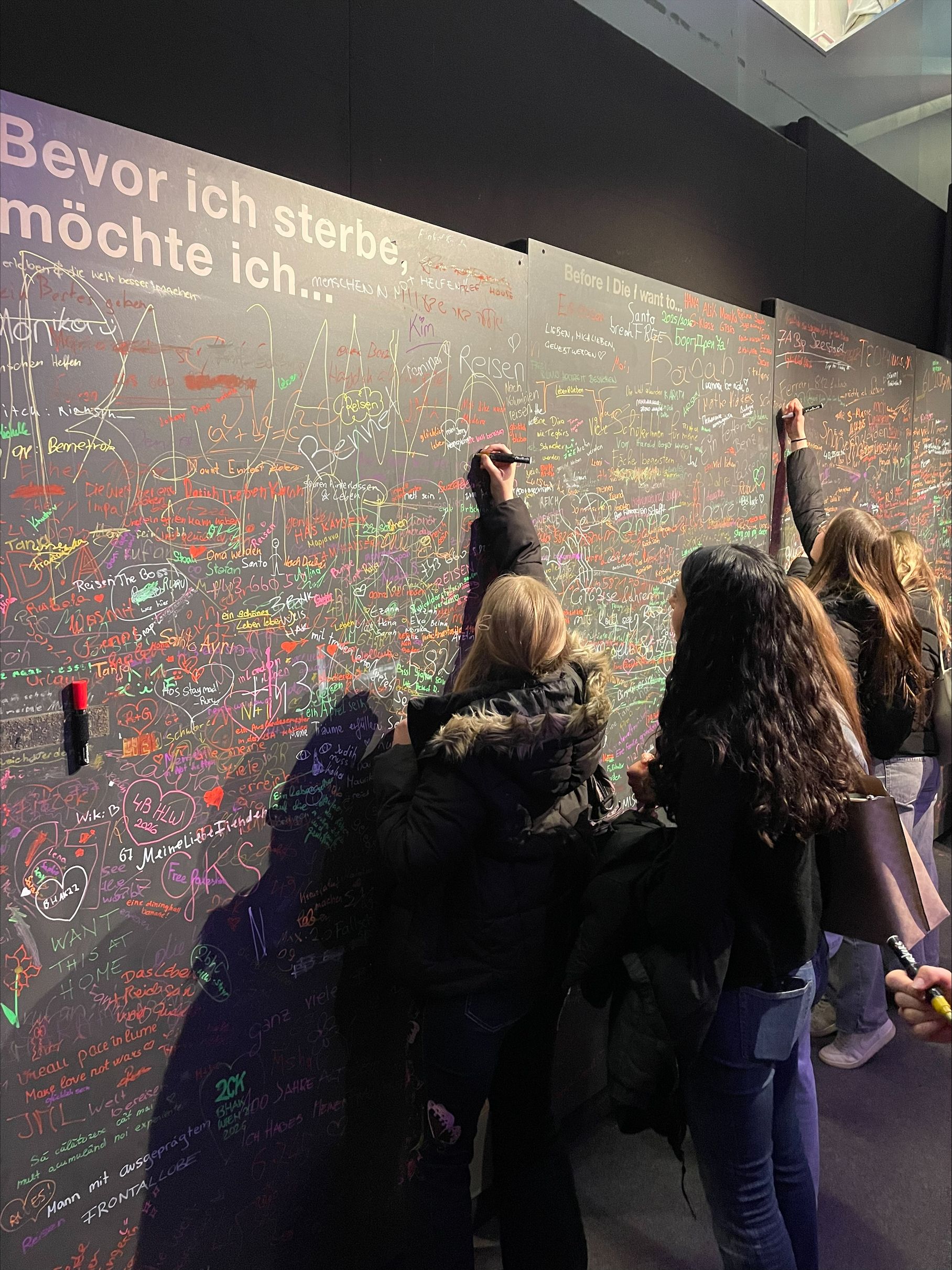 People writing on a colorful "Before I die, I want to..." wall filled with various messages and signatures in a public space.