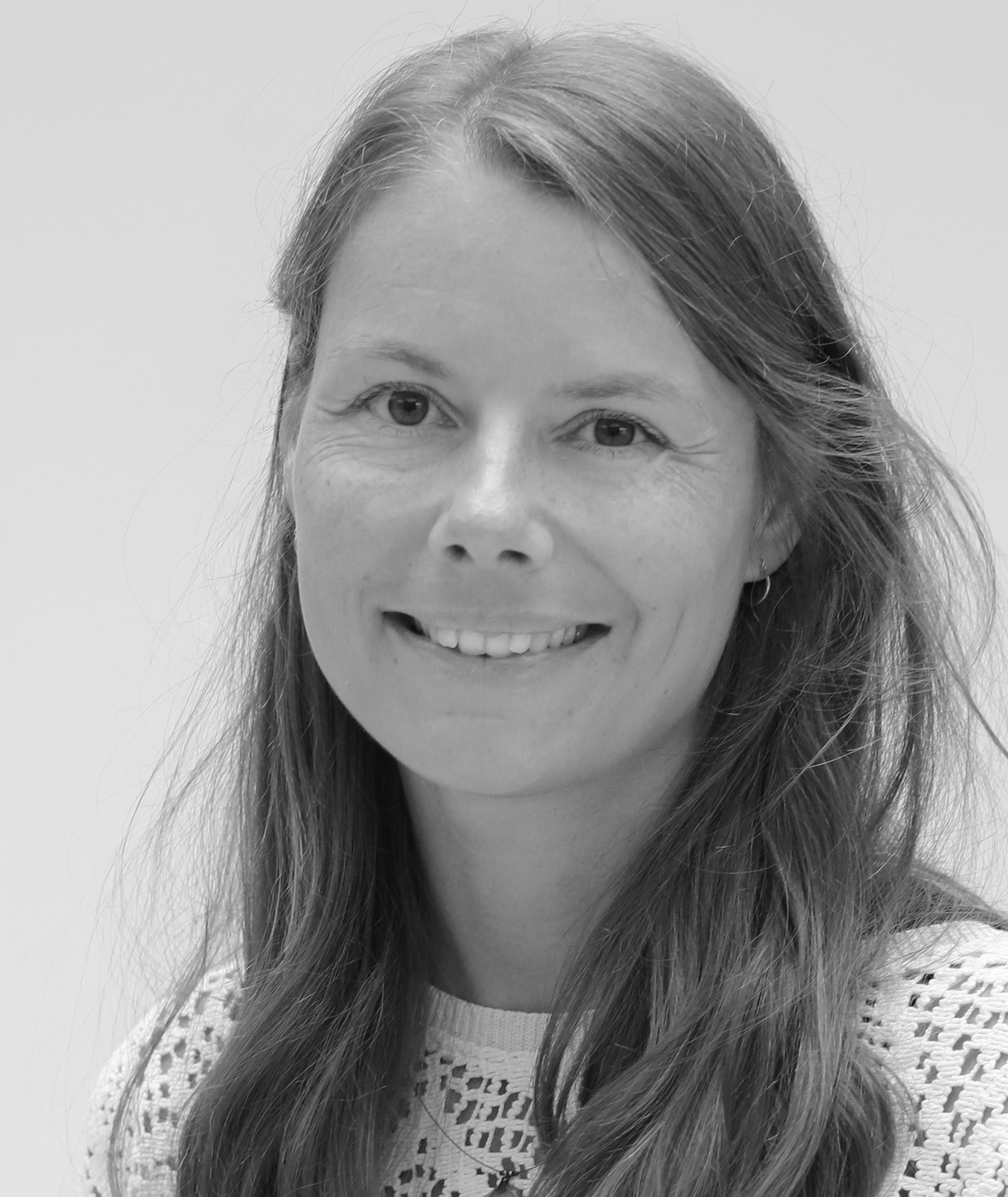 Smiling woman with long hair wearing a patterned top, looking at the camera against a plain background. Black and white photo.