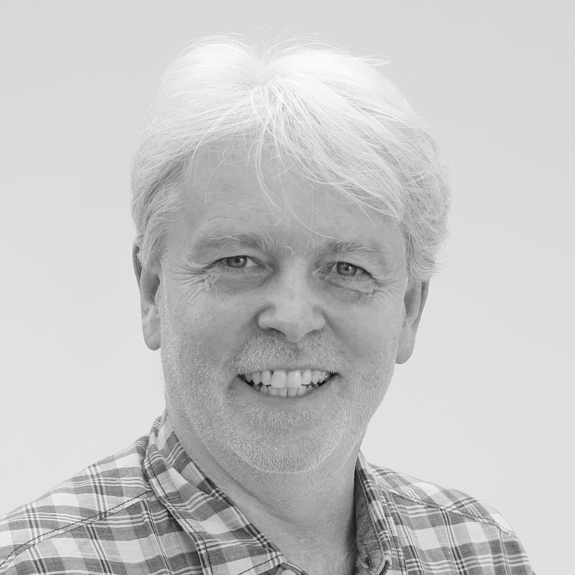 Smiling man with gray hair and a beard, wearing a plaid shirt, in a grayscale image.