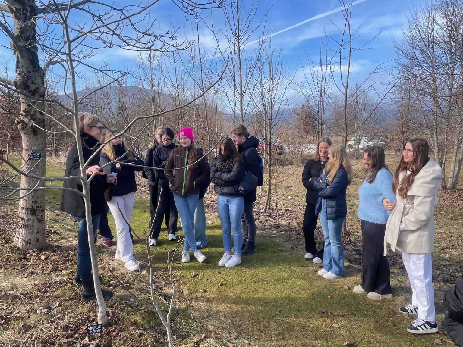 A group of people stand outdoors in a winter landscape, listening to a guide near leafless trees under a clear blue sky.
