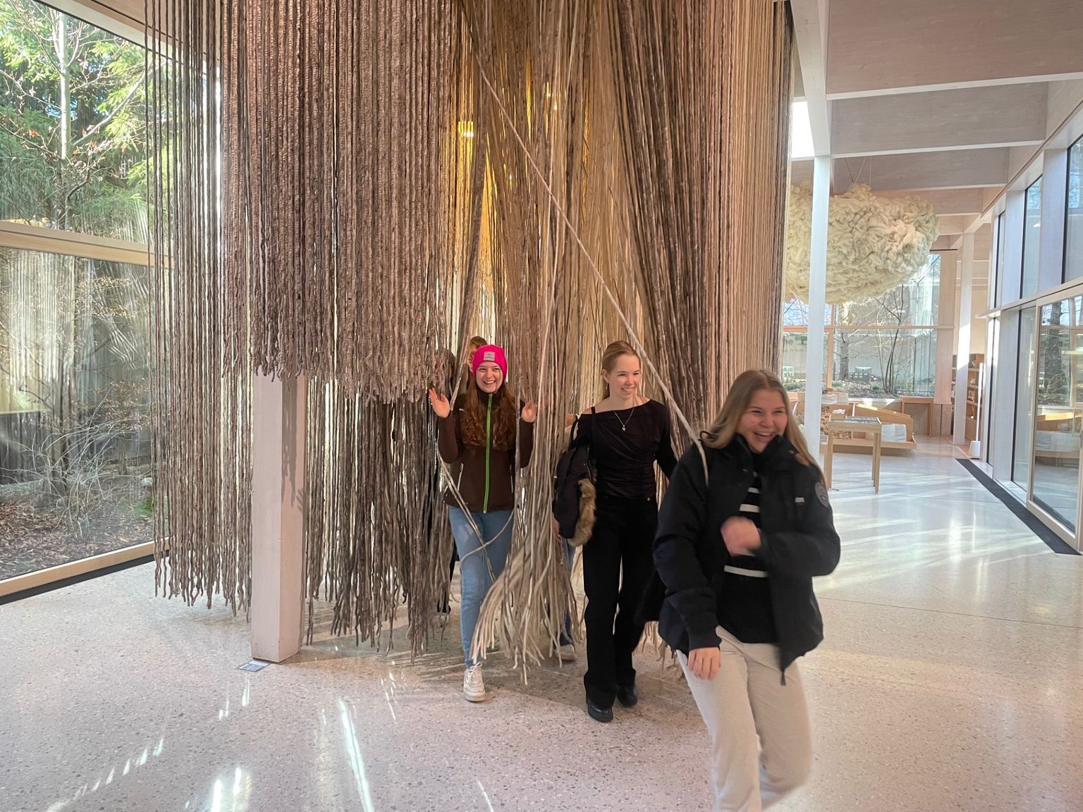 Three people walk through hanging strings in an art installation, smiling and enjoying the interactive exhibit in a sunlit gallery space.
