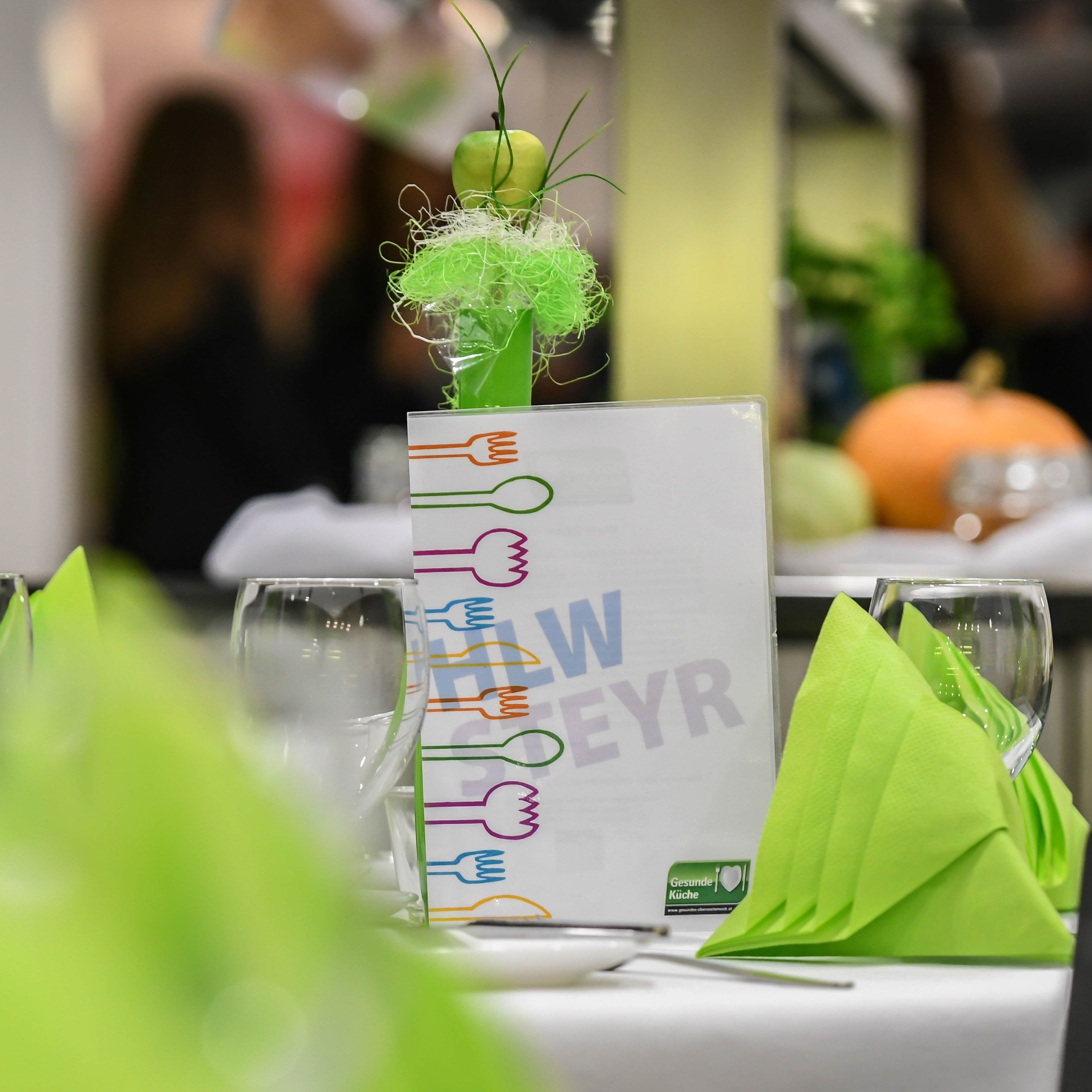 A table setting with green napkins, wine glasses, and a menu featuring colorful fork illustrations. A decorative apple is on top.