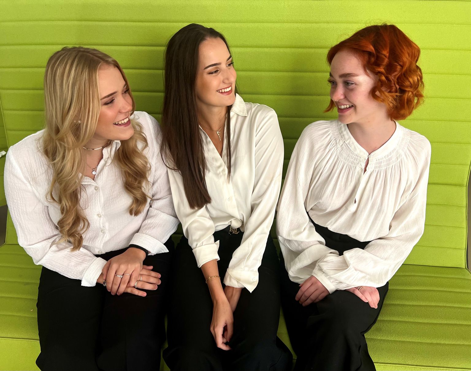 Three women in white blouses and black pants sit on a green bench, smiling and looking at each other.