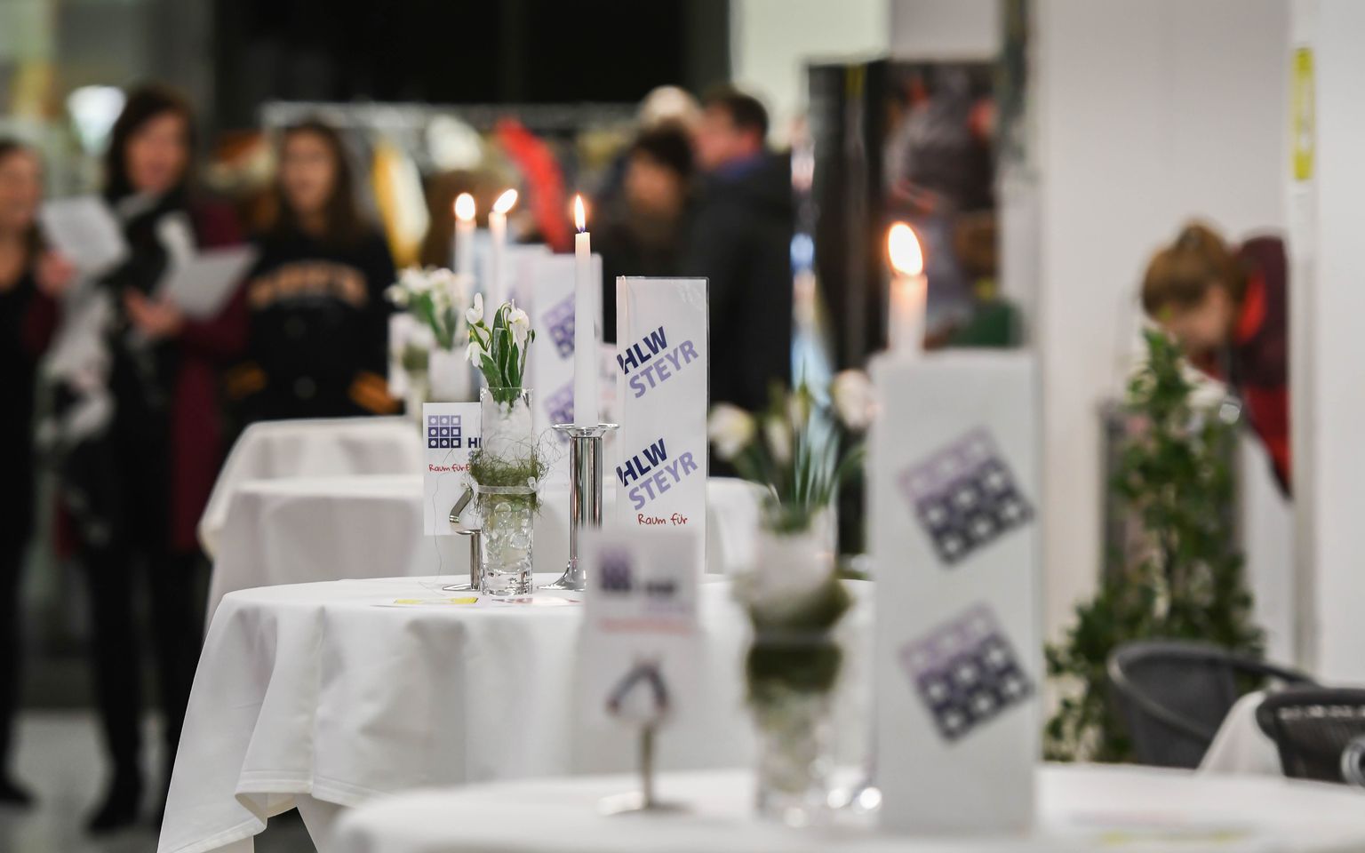 Tables with white tablecloths, candles, and flower arrangements are set up in a busy event space with people in the background.