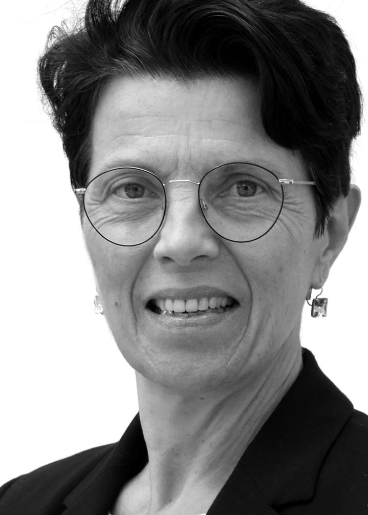 A person with short dark hair, wearing round glasses and a black jacket, smiles at the camera in a black and white portrait.