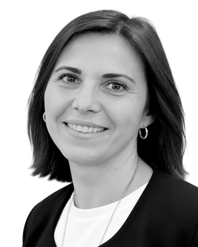 A black and white portrait of a woman with shoulder-length hair, wearing hoop earrings and a necklace, smiling at the camera.