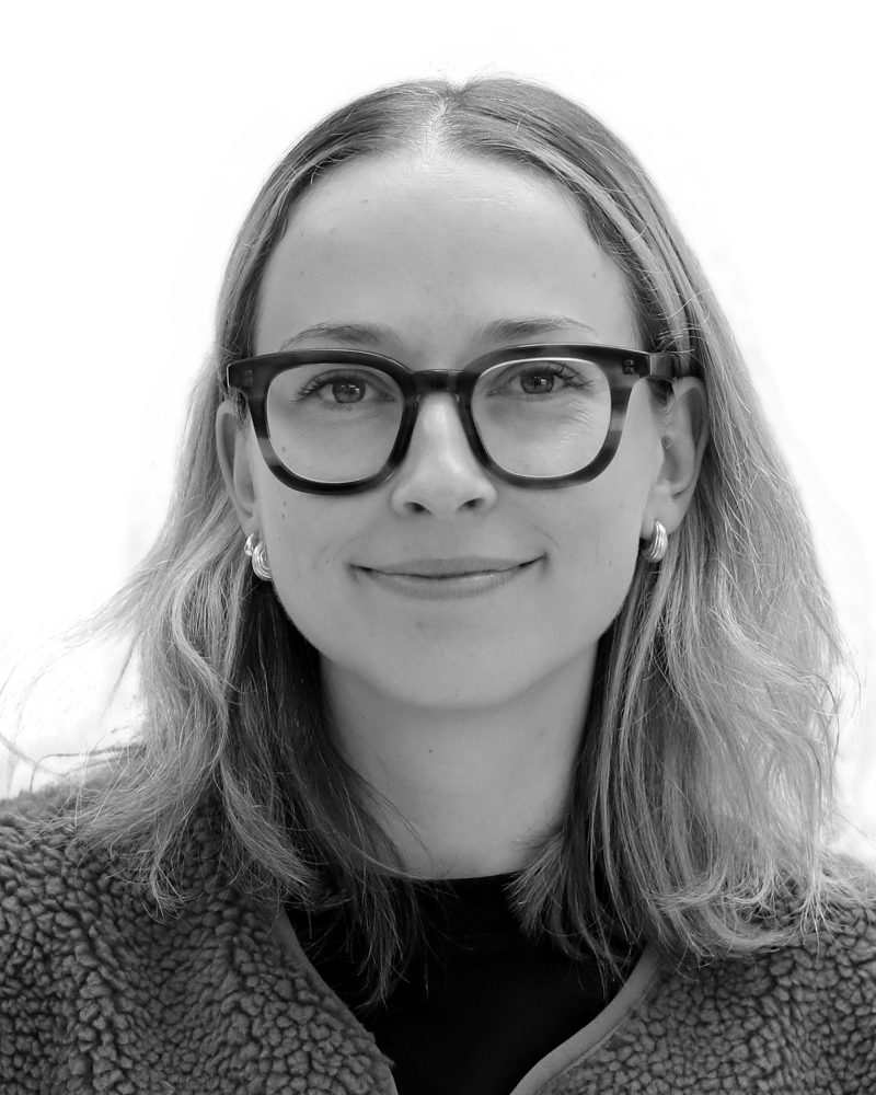 A woman with glasses and shoulder-length hair, wearing earrings and a textured jacket, smiles slightly in a black and white close-up photo.