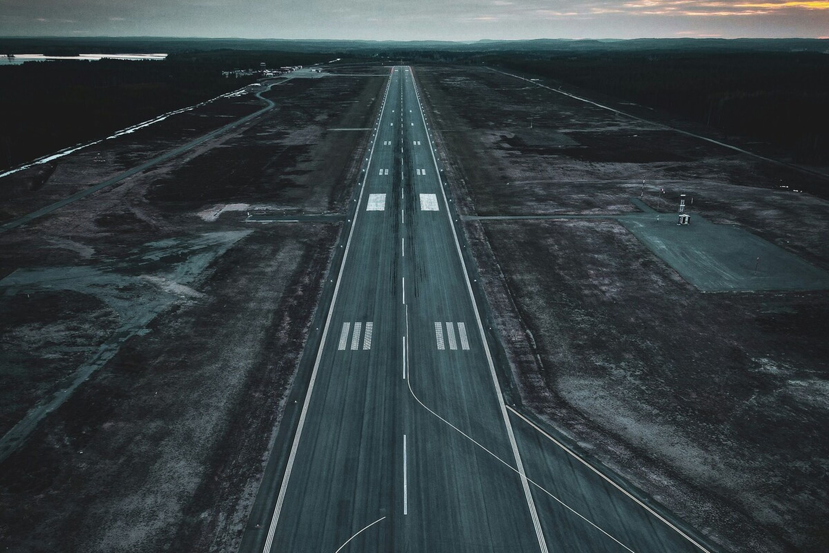 Luftaufnahme einer leeren Landebahn bei Sonnenuntergang, umgeben von karger Landschaft und dunklem Himmel.