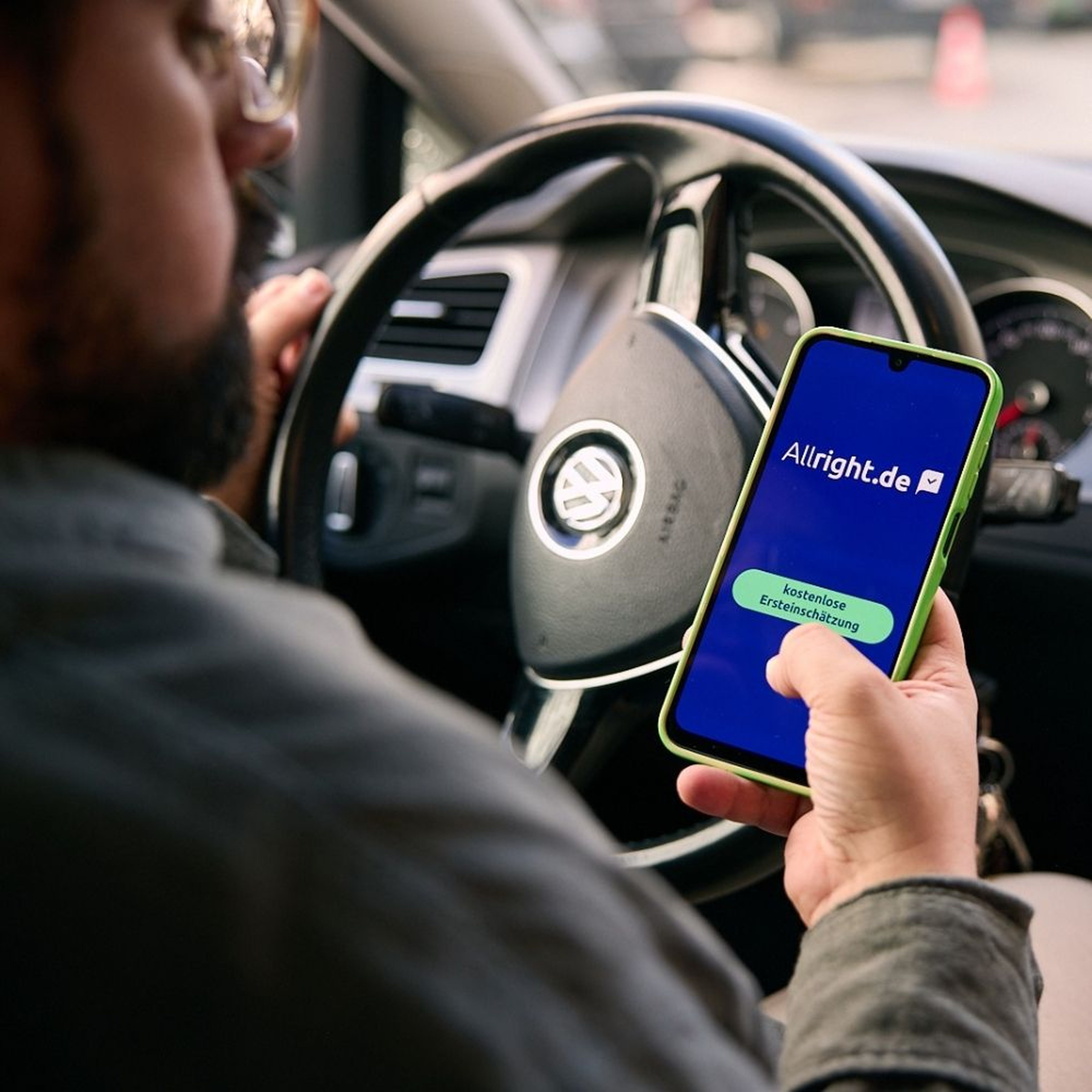 Eine Person in einem Auto mit einem Volkswagen-Lenkrad hält ein Smartphone in der Hand, auf dem die App „Allright.de“ zu sehen ist.
