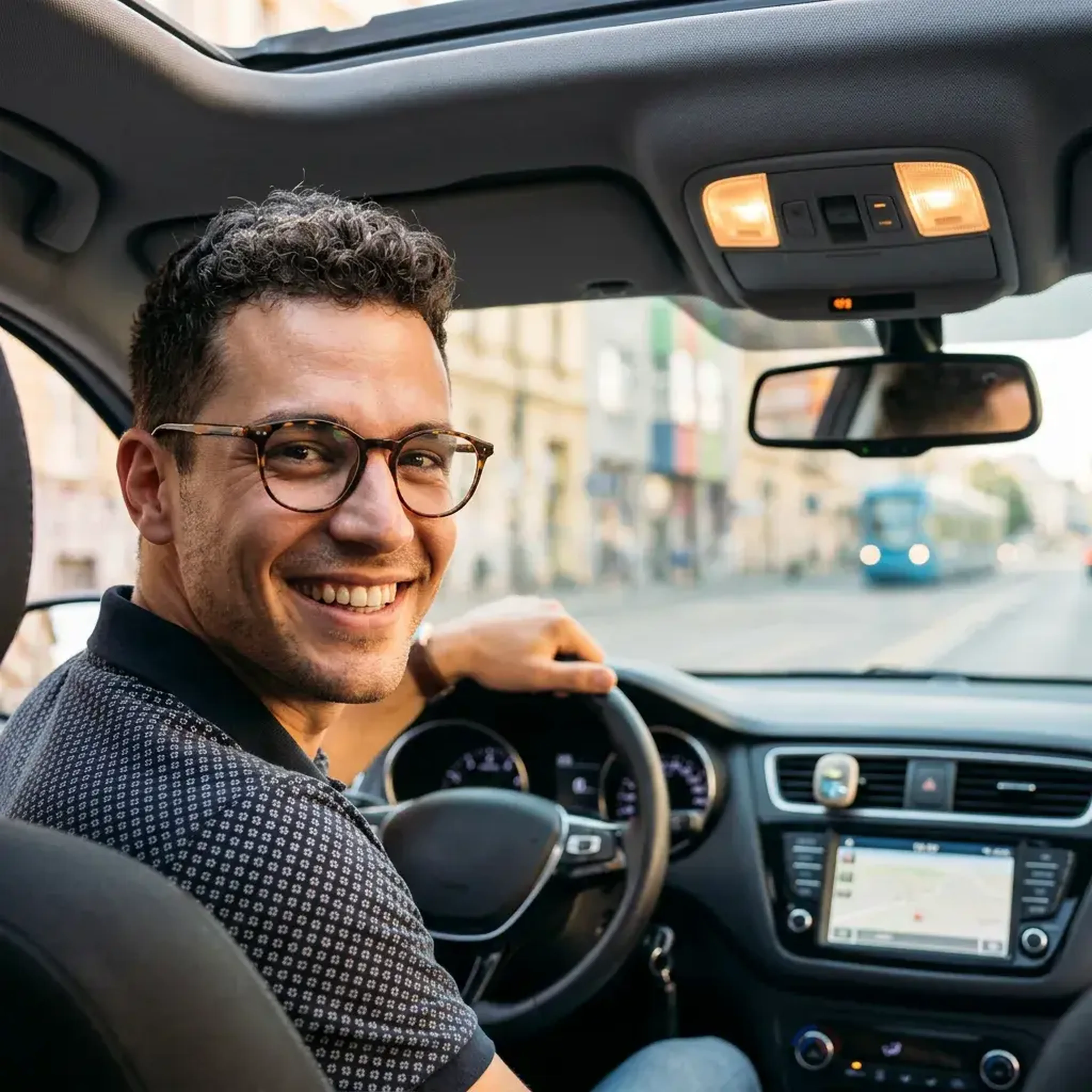 Ein Mann mit Brille sitzt lächelnd am Steuer eines Autos und blickt in Richtung Kamera. Durch die Windschutzscheibe ist eine Straße zu sehen.