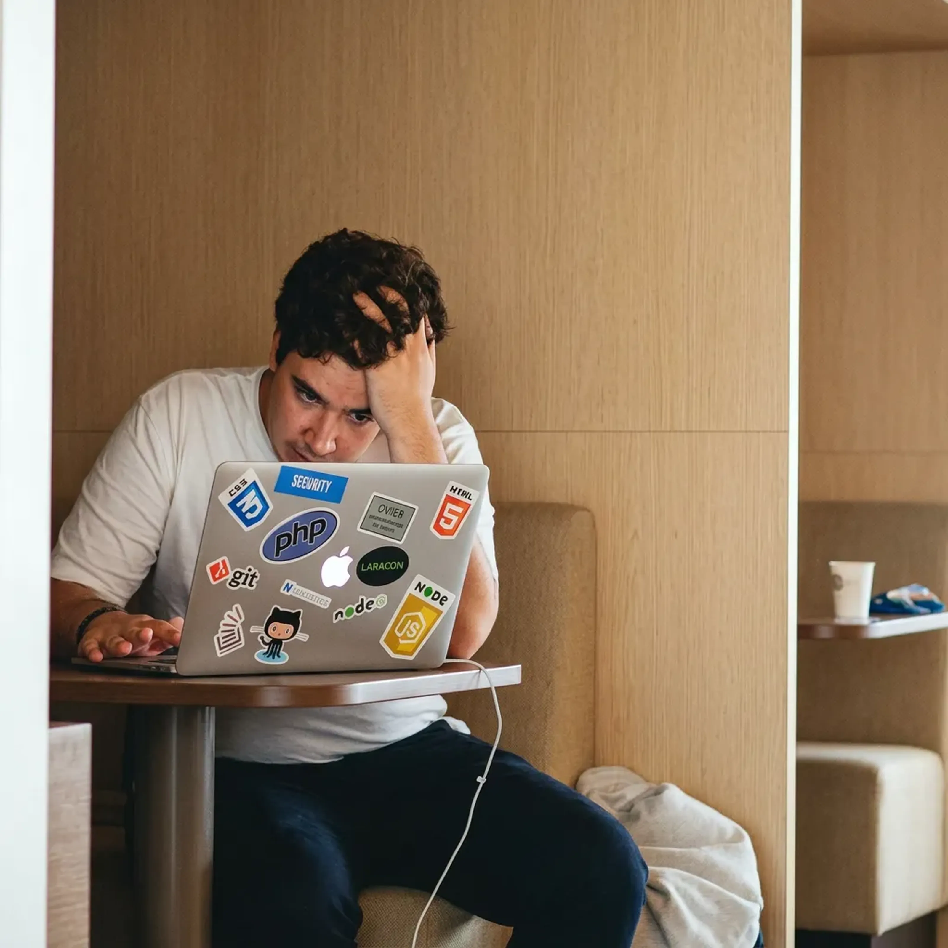 Junger Mann in einem weißen T-Shirt sitzt konzentriert vor einem Laptop mit Programmieraufklebern an einem Tisch in einem modernen Innenraum.