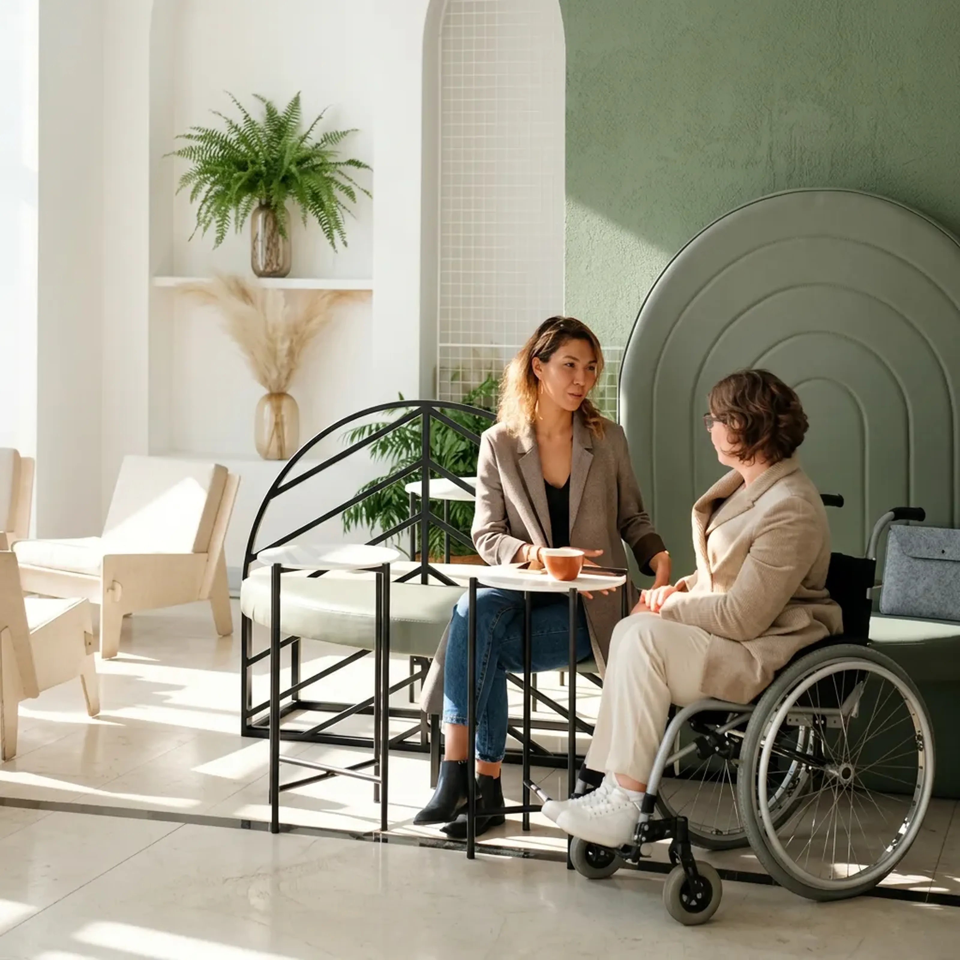 Zwei Frauen in einem Café, eine im Rollstuhl, unterhalten sich an einem Tisch. Die Umgebung ist modern und grün dekoriert.