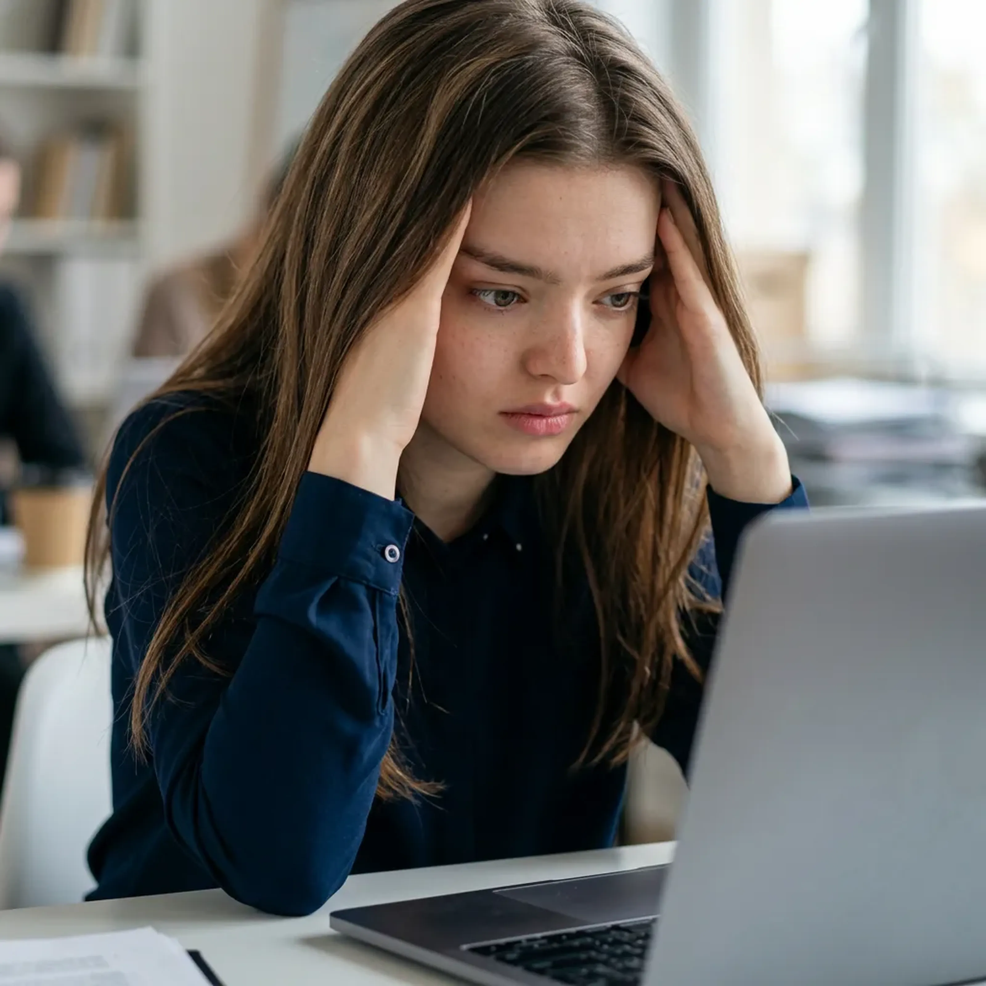 Eine Frau sitzt mit nachdenklichem Gesichtsausdruck vor einem Laptop und stützt ihren Kopf mit beiden Händen.