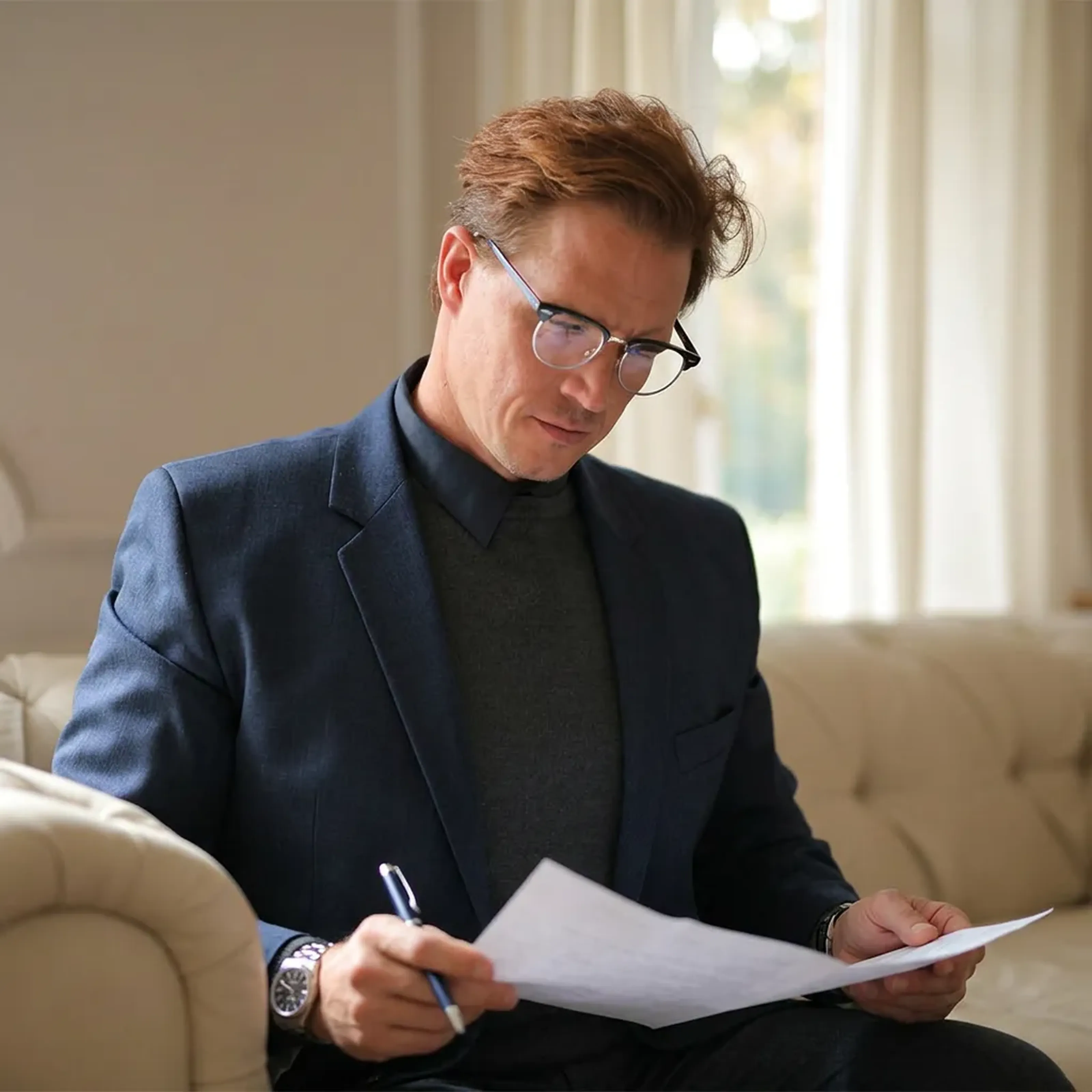 Mann in Anzug liest konzentriert Dokumente, sitzt auf einem beigen Sofa, hält einen Stift in der Hand, helle Vorhänge im Hintergrund.