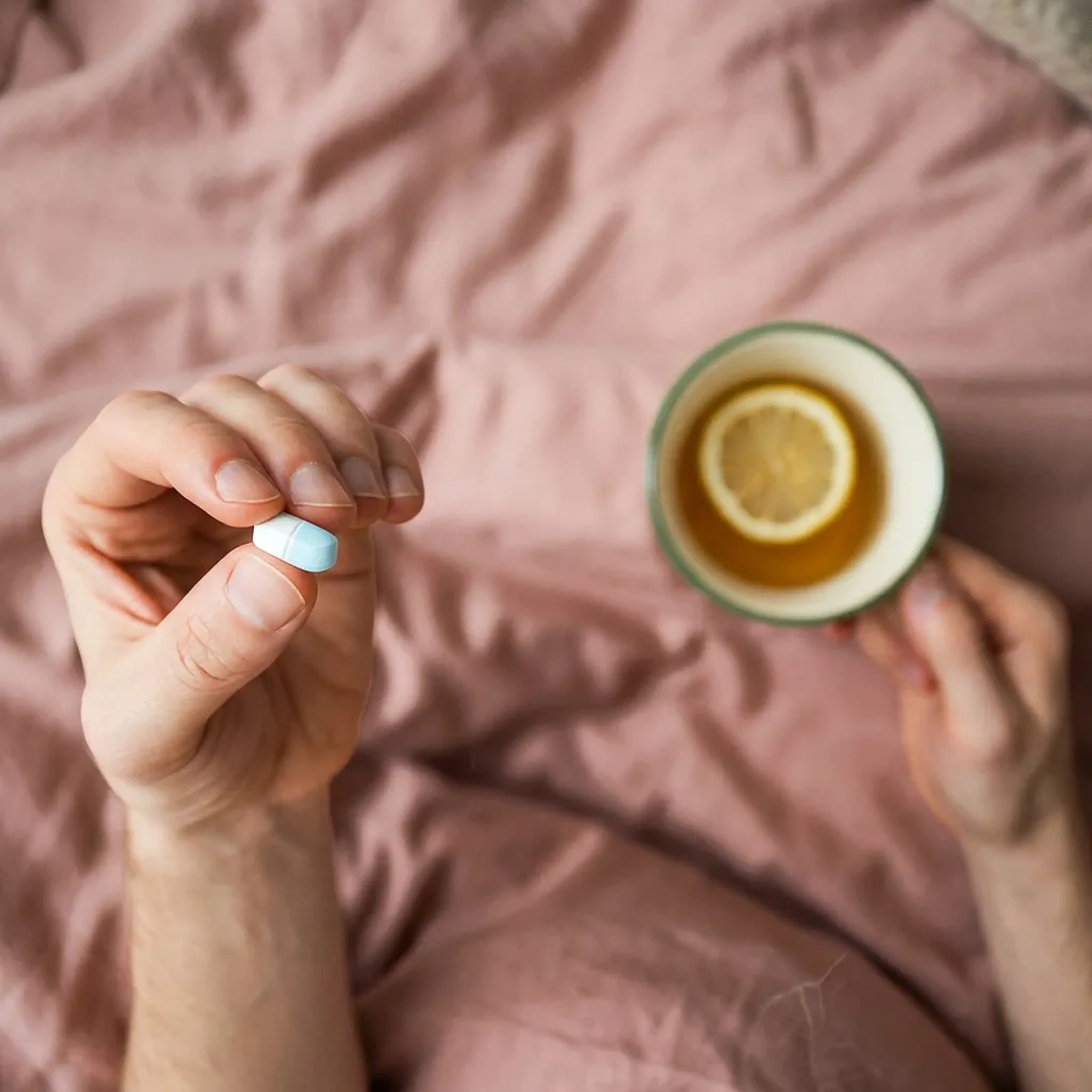 Hand hält eine blaue Tablette über einem Bett, während die andere Hand eine Tasse Tee mit Zitronenscheibe hält.