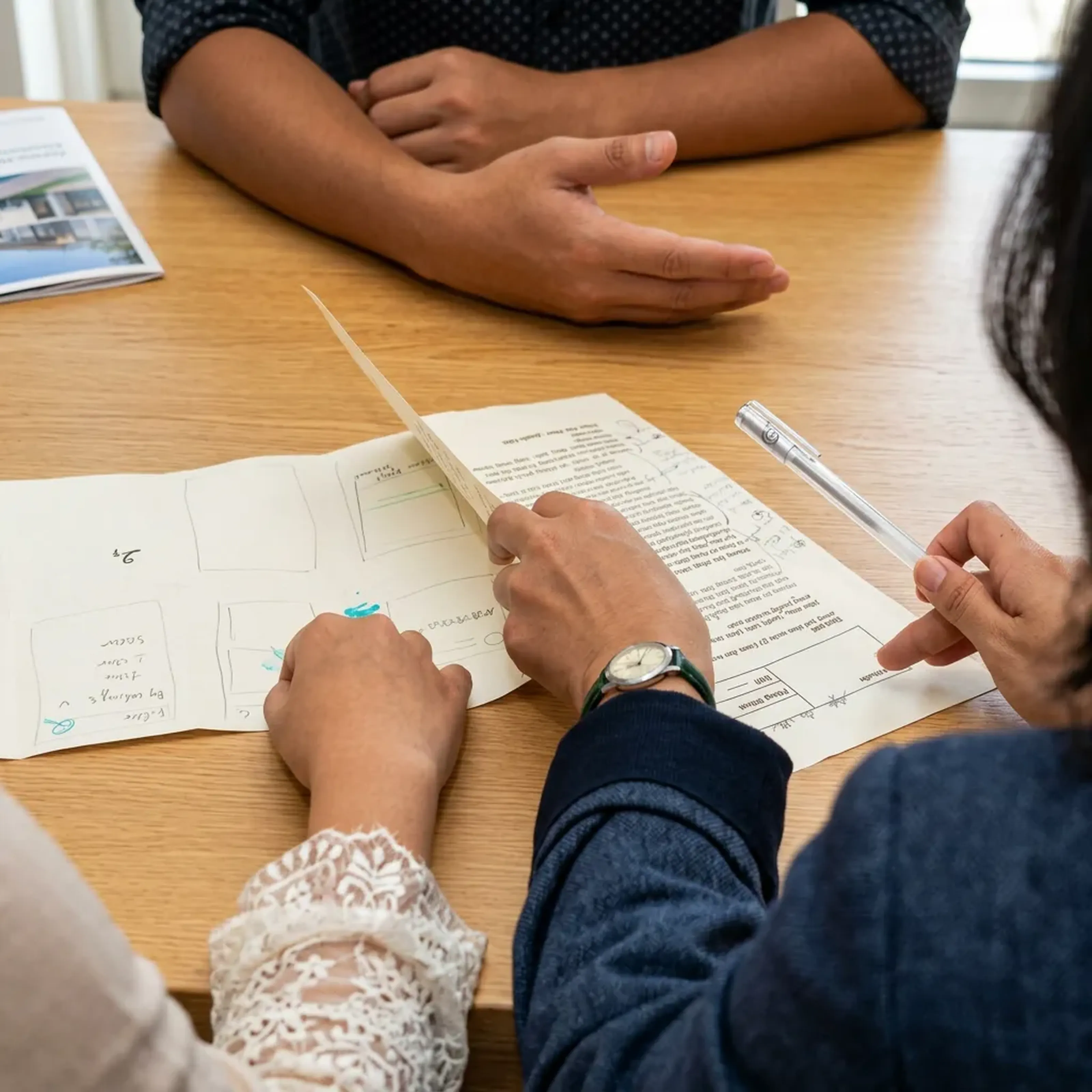 Drei Personen sitzen an einem Tisch und diskutieren über Papiere. Eine Person zeigt mit einem Stift auf das Dokument.