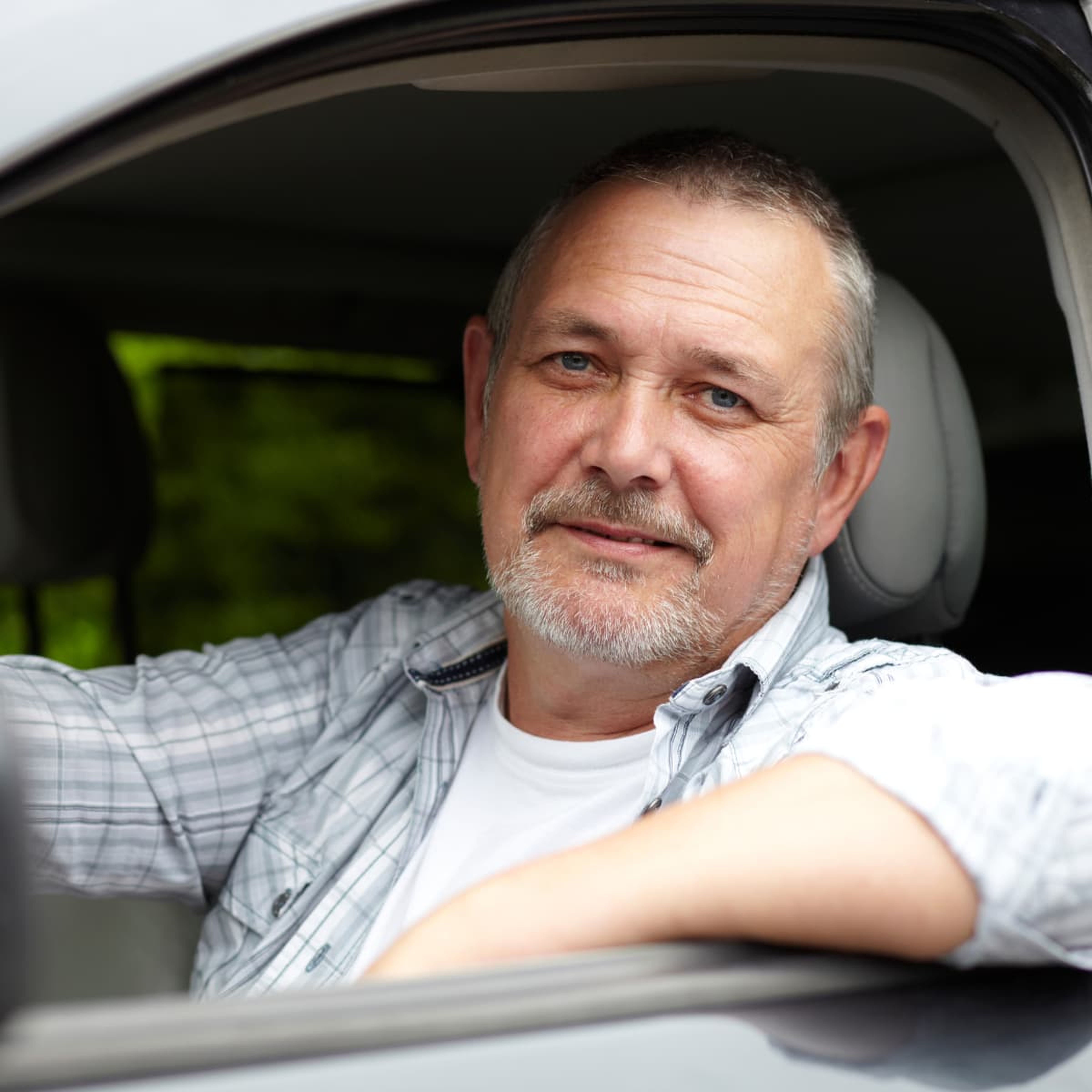 Mann mit kurzen Haaren und Bart, der lächelnd in einem Auto sitzt, ein kariertes Hemd trägt und vom Fahrersitz aus direkt in die Kamera blickt.