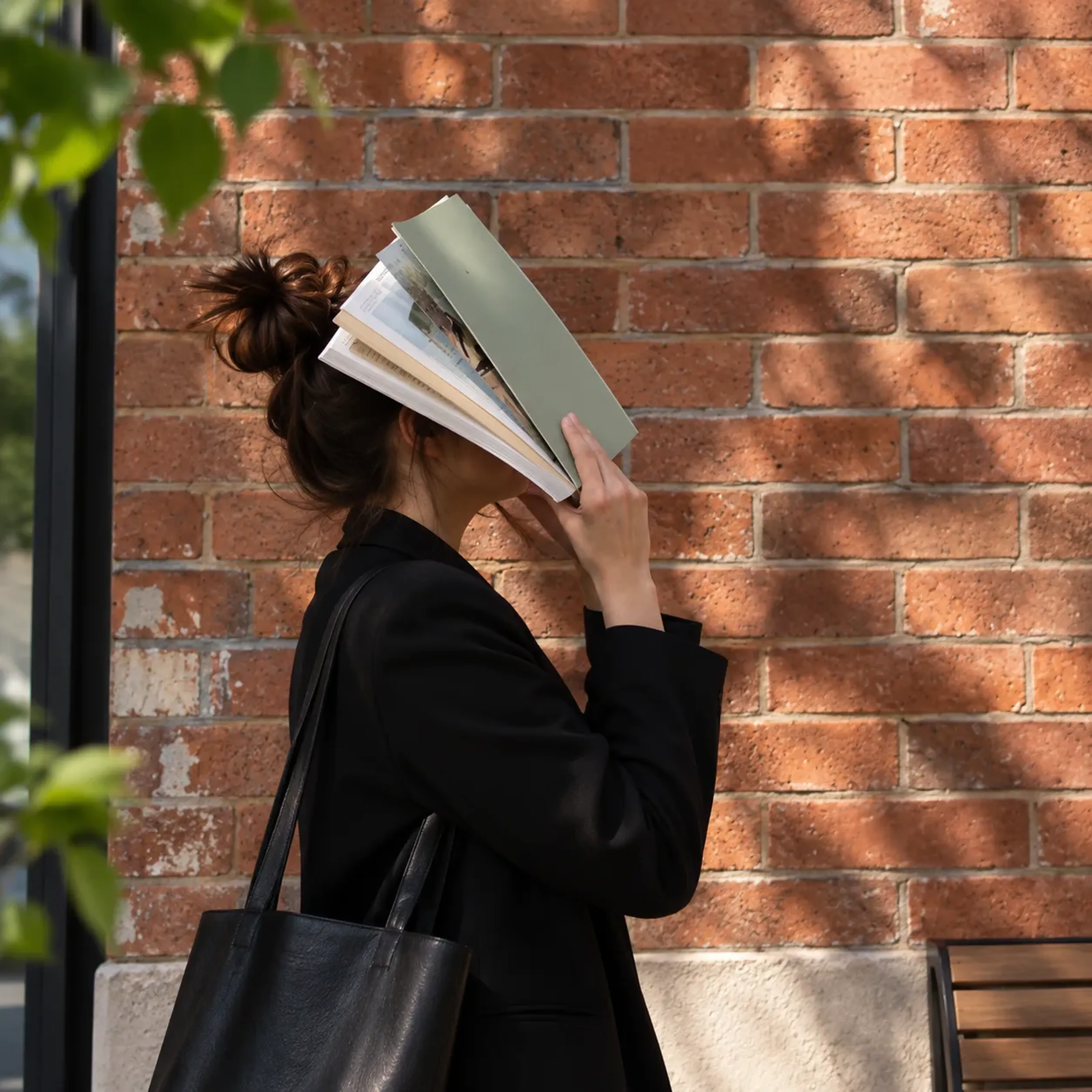Frau in schwarzem Blazer hält ein Buch vor ihr Gesicht, steht an einer Ziegelmauer. Sonnenlicht und Schatten fallen auf die Szene.