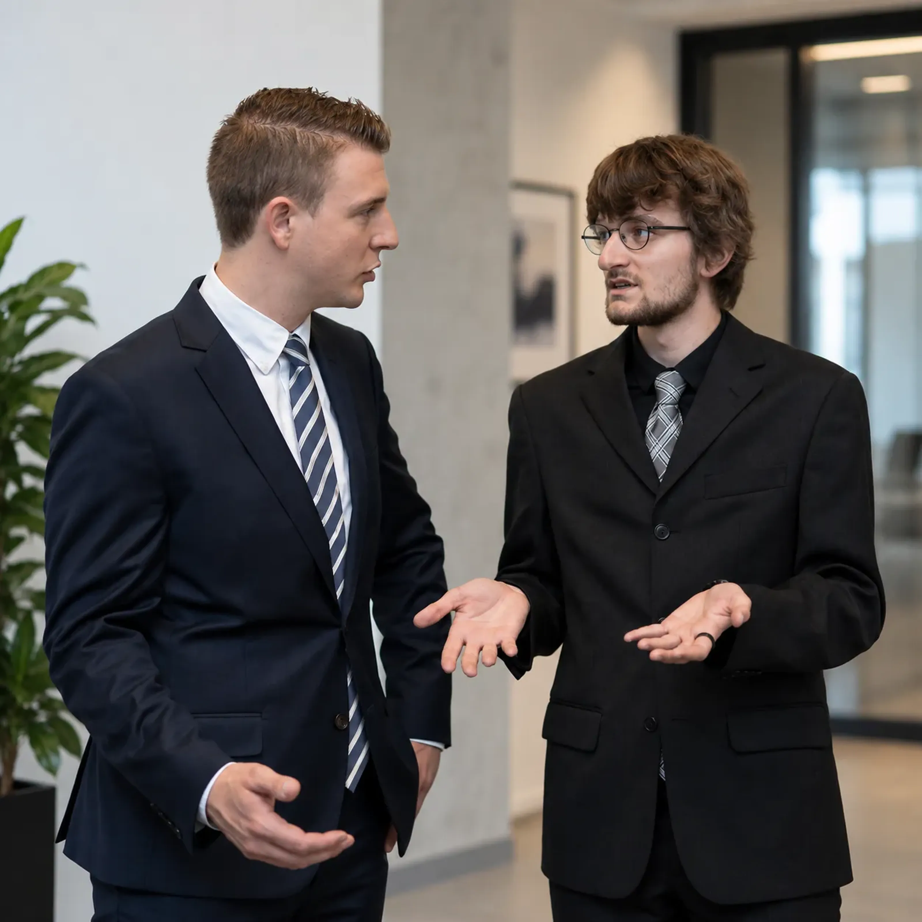 Zwei Männer in Anzügen unterhalten sich gestikulierend in einem modernen Büro mit Pflanzen im Hintergrund.