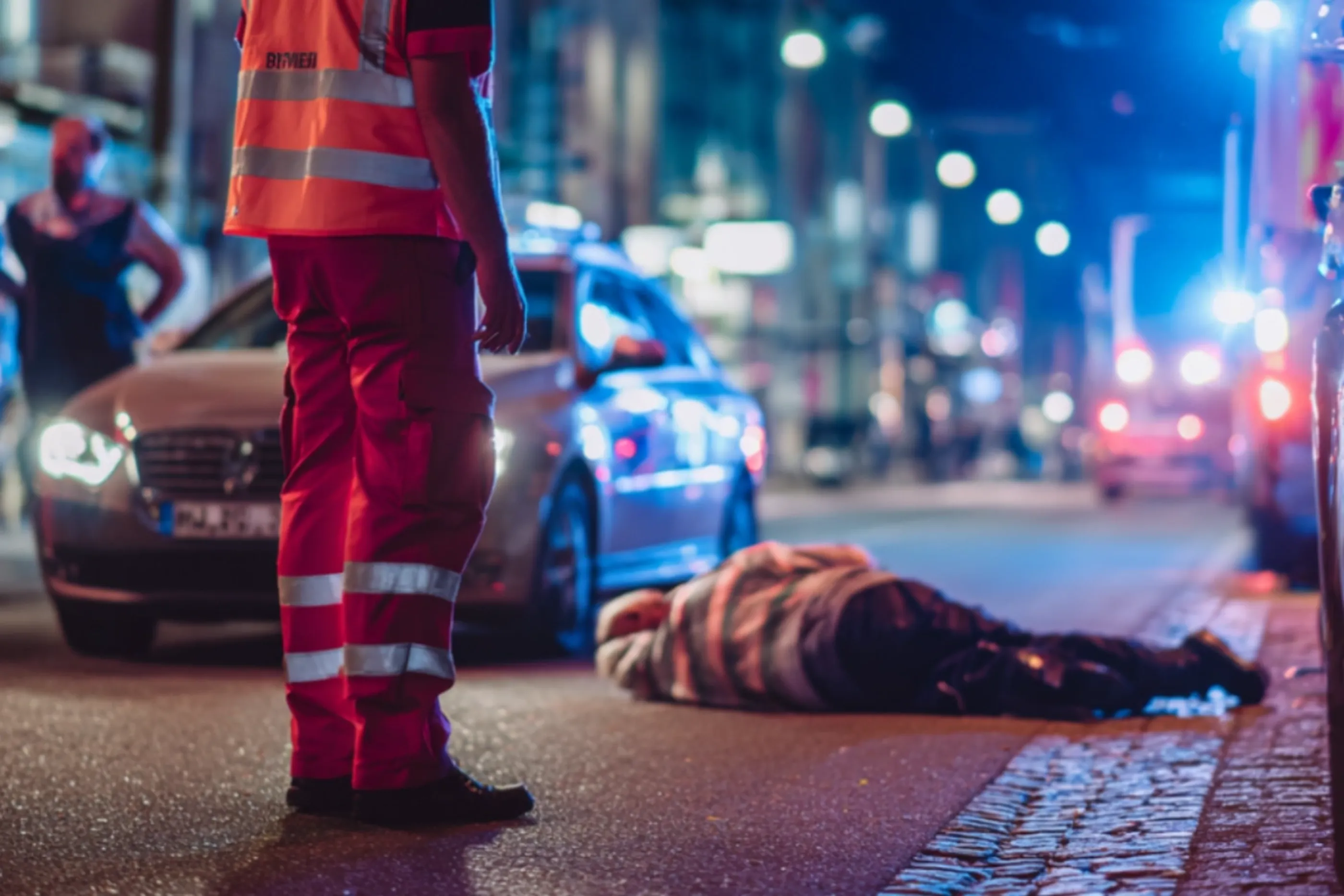 Person in Rettungsweste hilft einer Person auf der Straße bei Nacht, umgeben von Autos und Einsatzfahrzeugen.