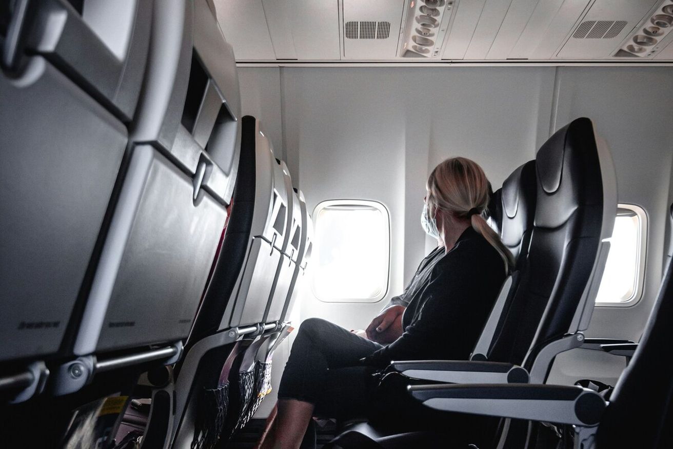 Frau mit Maske sitzt alleine in einem Flugzeug, schaut aus dem Fenster.