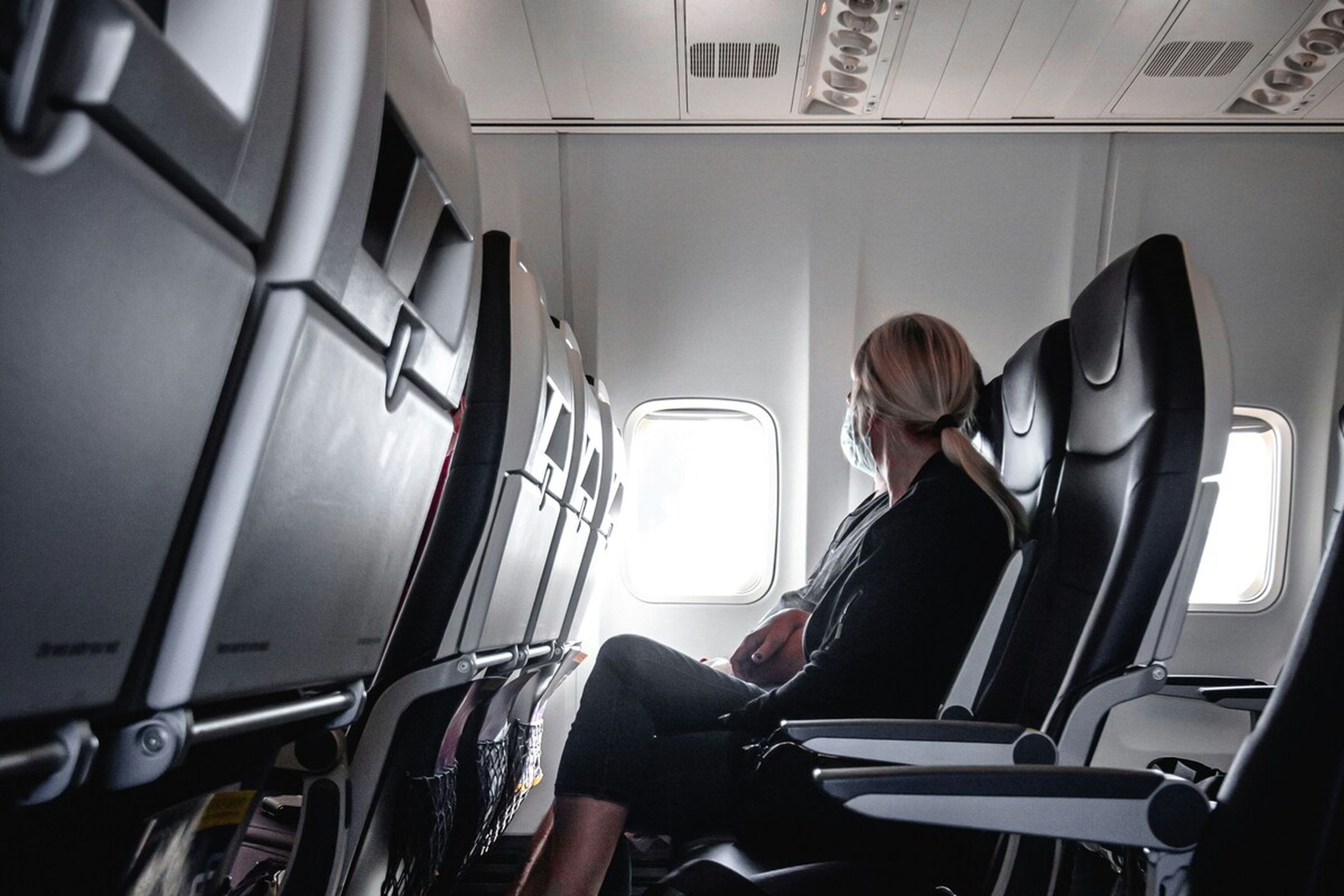 Frau mit Maske sitzt alleine in einem Flugzeug, schaut aus dem Fenster.