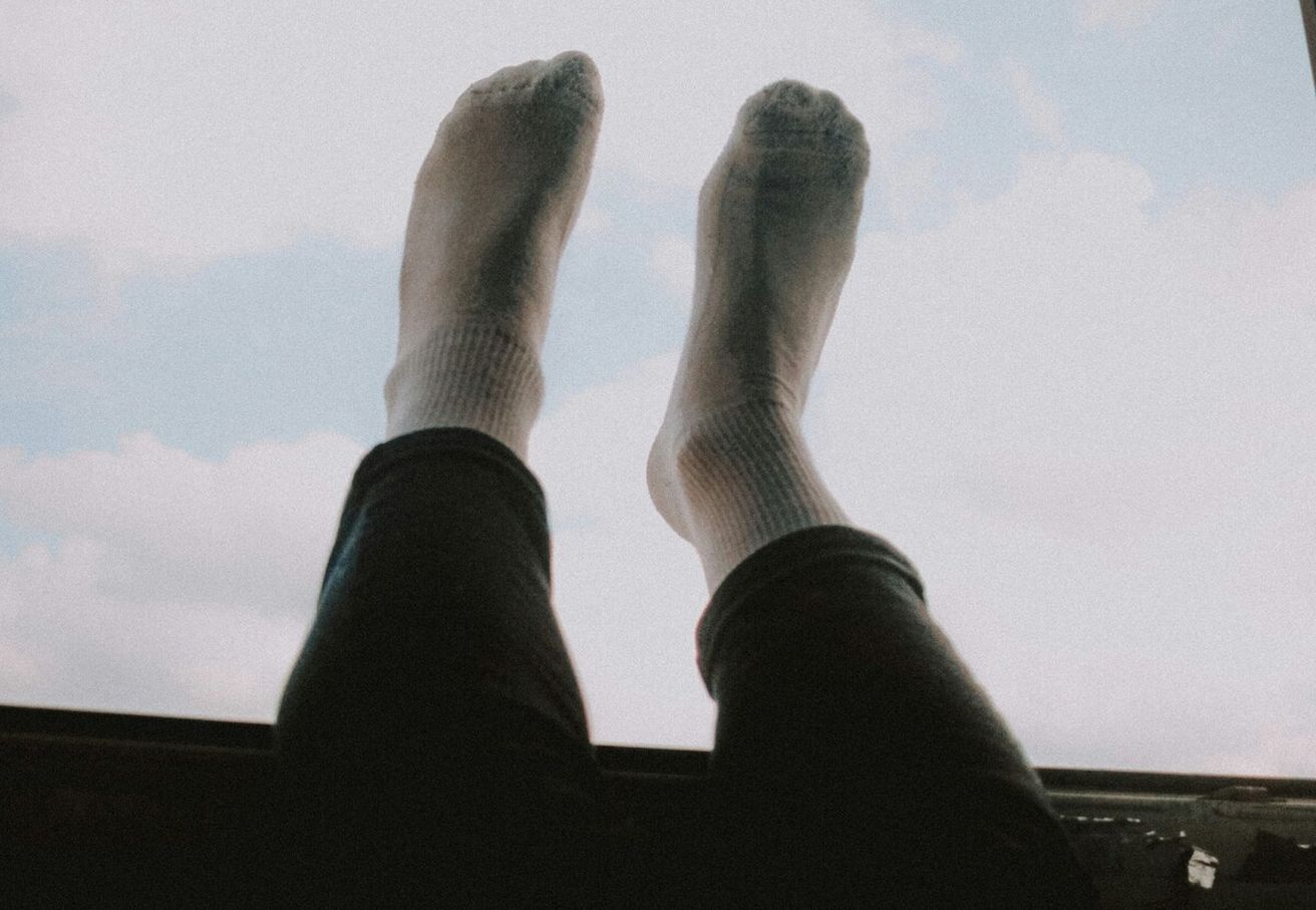 Person mit weißen Socken streckt Beine in den Himmel, getragen von dunkel gekleideten Beinen, vor blauem Himmel.