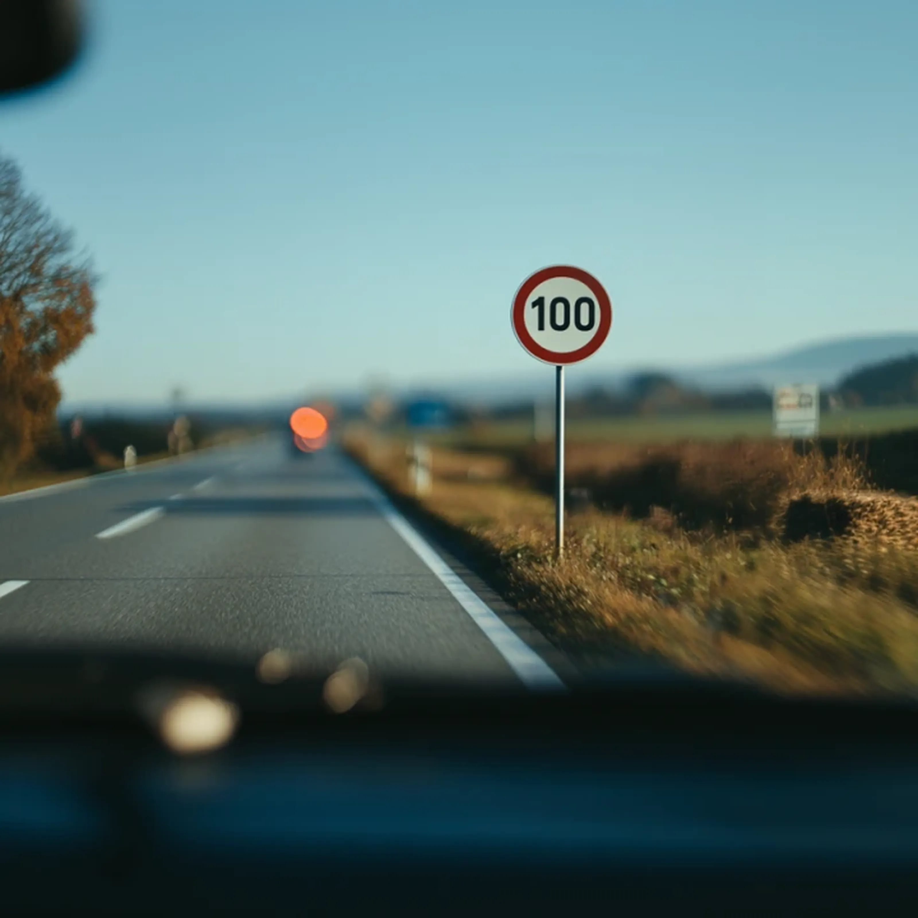 A rural road with a speed limit sign reading "100," fields and trees on the side, and hills visible in the background.