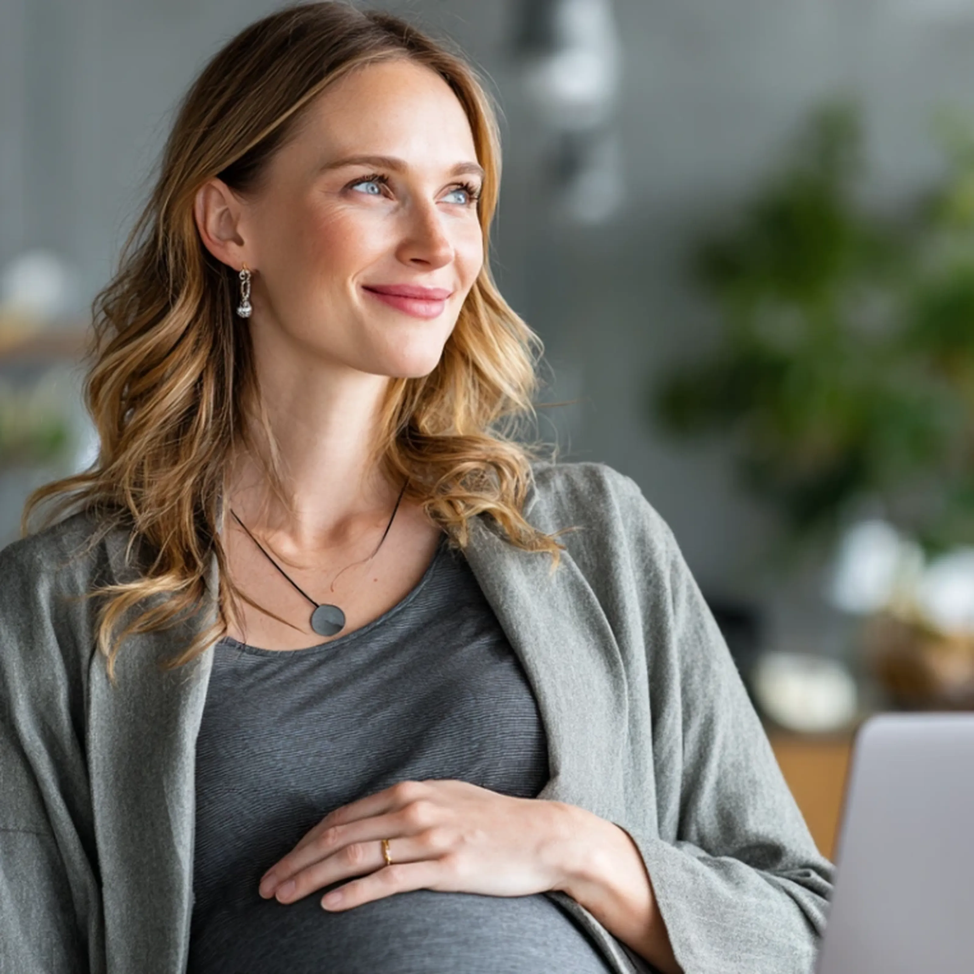 Smiling pregnant woman in a gray blazer and top, touching her belly, looking out thoughtfully in a softly lit, blurred interior setting.