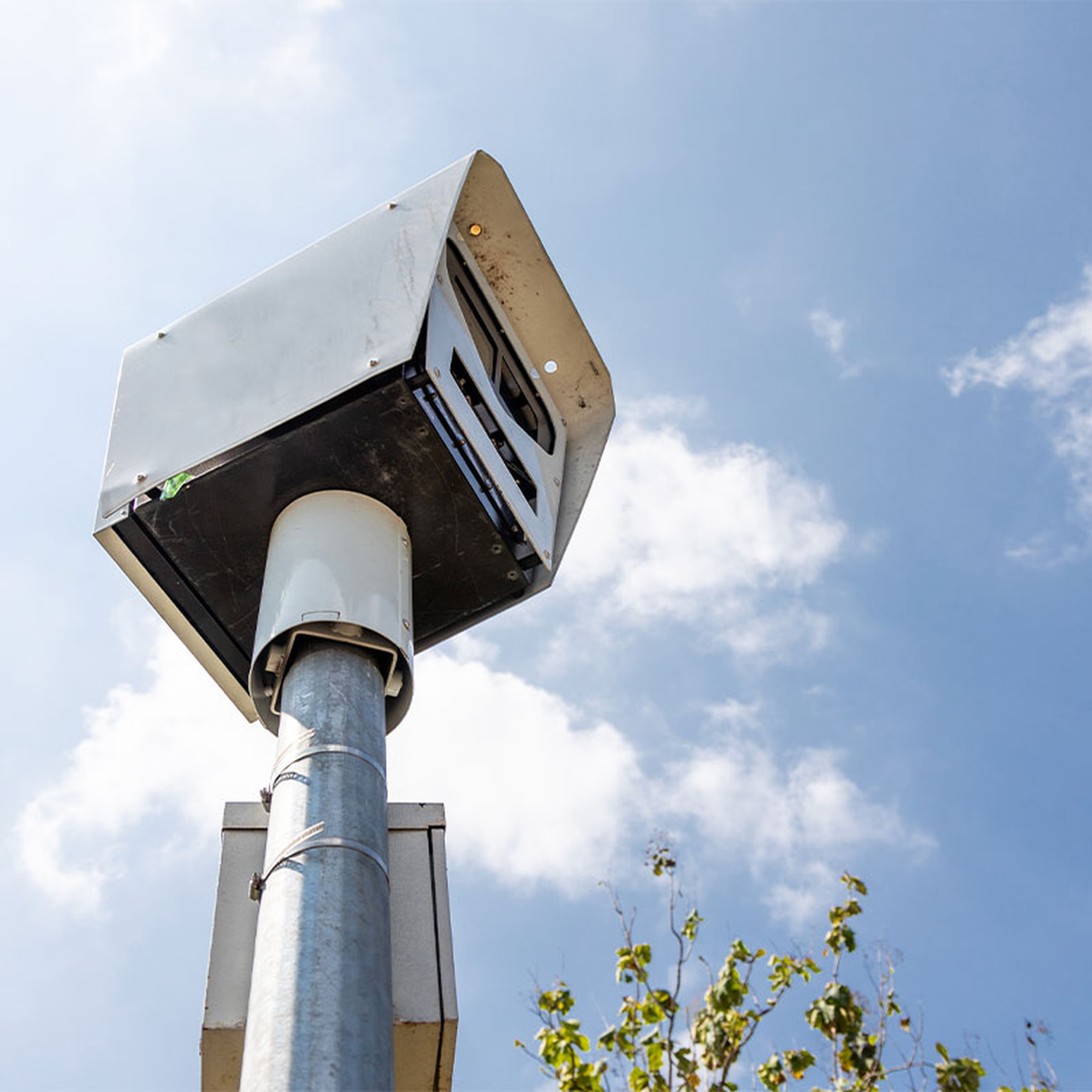 Pole-mounted speed camera against a blue sky with scattered clouds and some tree branches partially visible at the bottom.