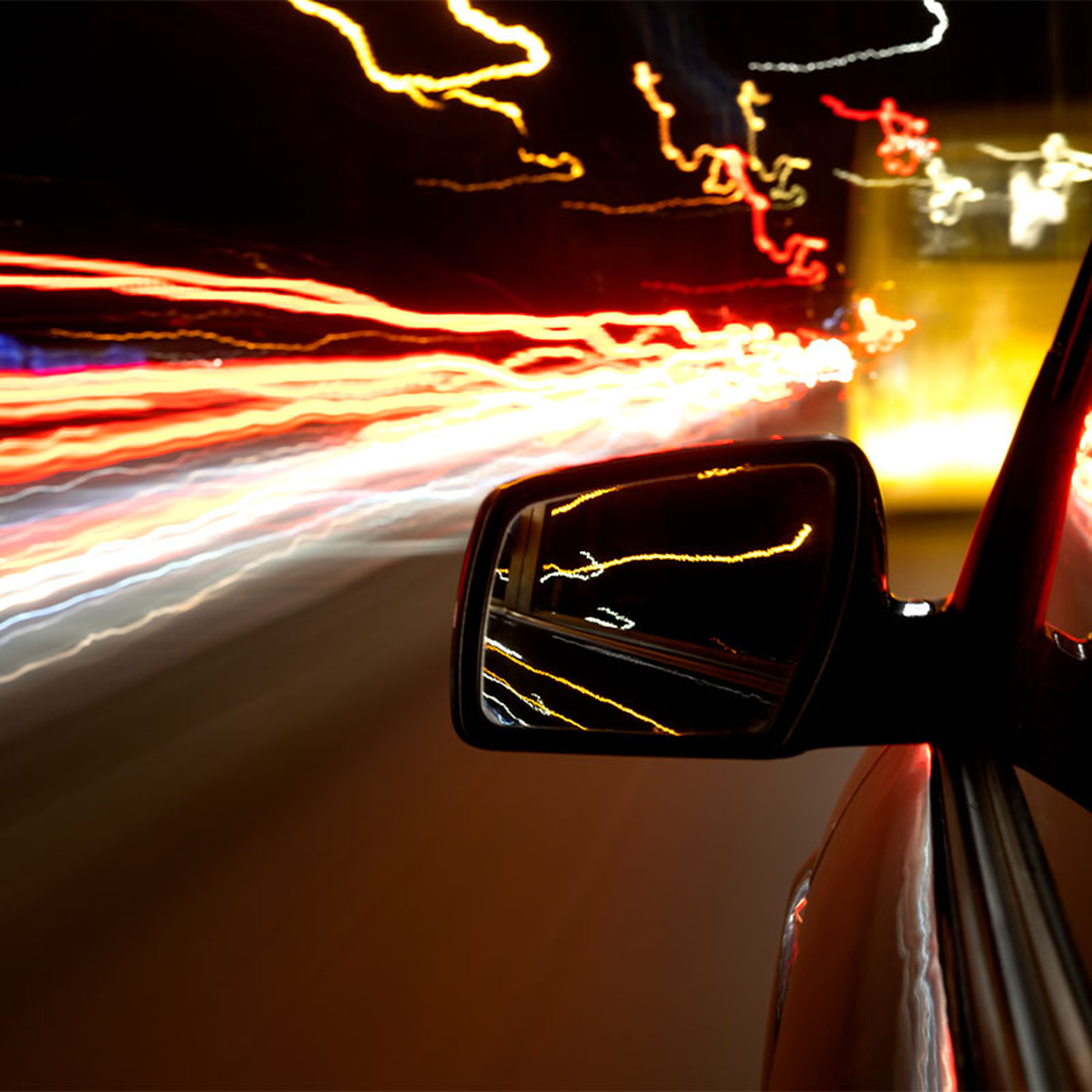 Car side mirror showing motion blur of colorful streaks of light, creating a dynamic and fast-paced night driving scene.