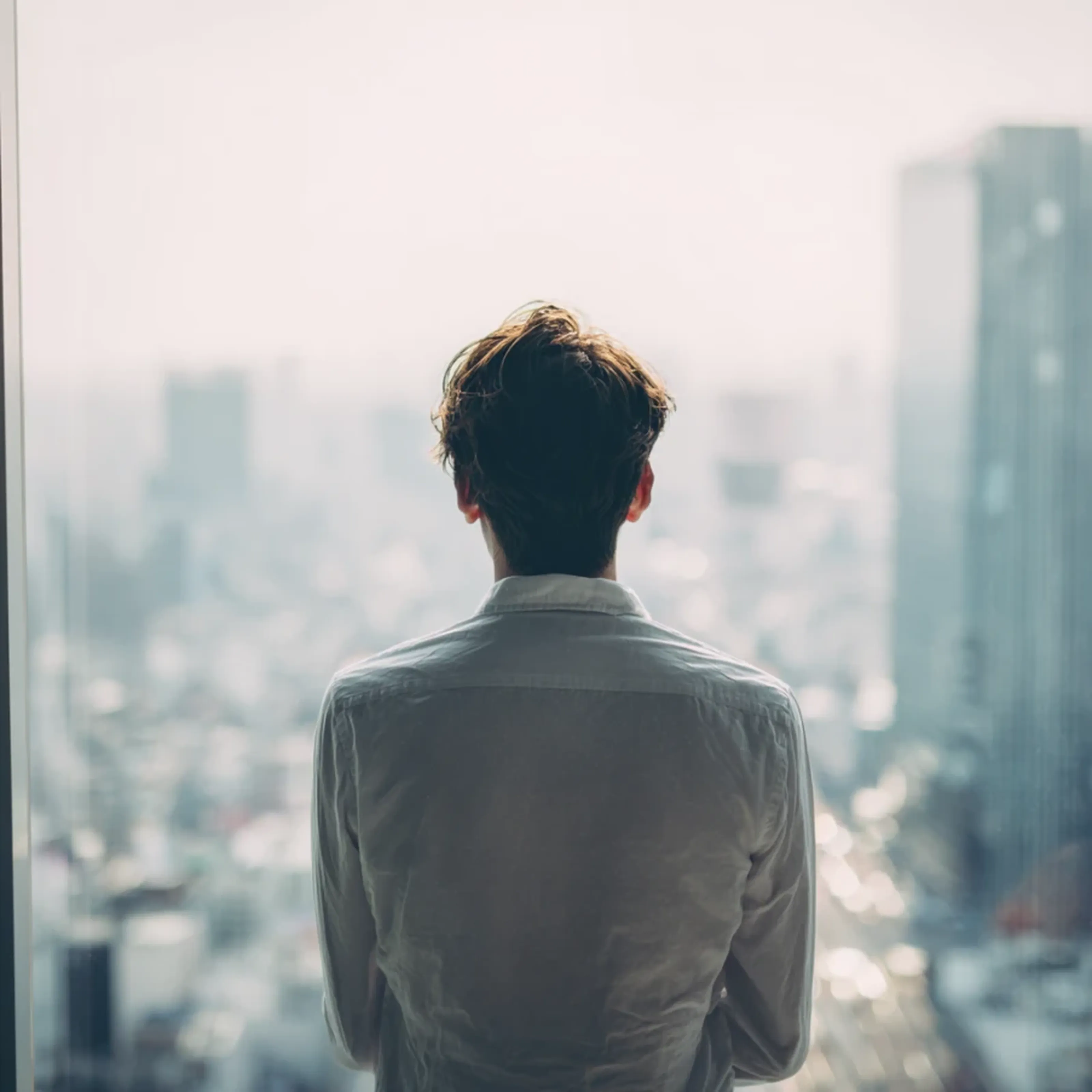 A person in a white shirt looks out at a cityscape through a large window, with skyscrapers in the background.