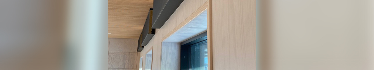 Black rectangular ventilation ducts on a wood-paneled ceiling above windows in a light-colored wall