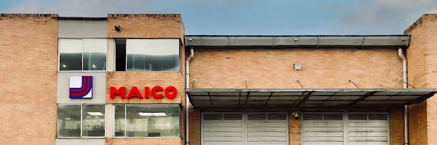 Fassade eines Backsteingebäudes mit großen Fenstern, zwei geschlossenen Rolltoren und rotem Schriftzug 'MAICO' neben einem Logo mit blauen und roten Linien.