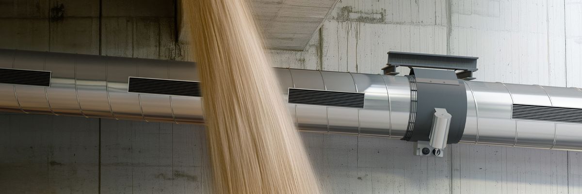 Großer silberner Industrieventilator an einer Betonwand, vor dem ein großer Strahl von feinem Material nach unten fällt.