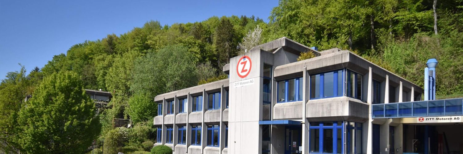 Mehrstöckiges Bürogebäude mit blauen Fenstern und Zitt Logo, davor geparkte weiße Fahrzeuge, umgeben von grünen Bäumen.