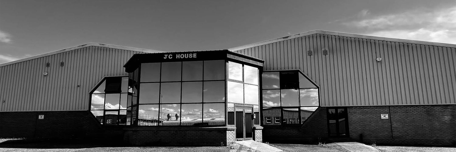 Großes Industriegebäude mit dreieckigem Glasvorbau und Schriftzug JC House und reflektierenden Fenstern unter bewölktem Himmel.