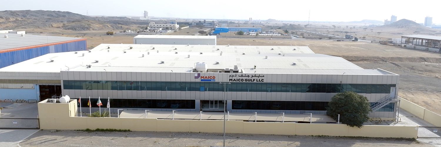 Flaches, rechteckiges Industriegebäude mit weißem Dach und MAICO-Gulf-Logo an der Fassade, umgeben von karger Landschaft.