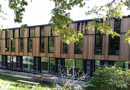 Two-story building facade with large windows and vertical wooden panels surrounded by greenery