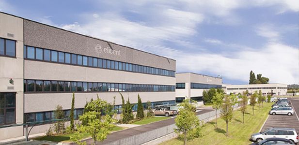 Großes, modernes Bürogebäude mit mehreren Fenstern und Schriftzug 'elicent' davor Bäume und geparkte Autos.