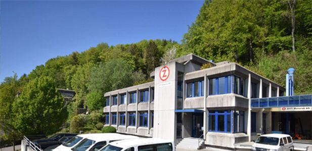 Mehrstöckiges Bürogebäude mit blauen Fenstern und Zitt Logo, davor geparkte weiße Fahrzeuge, umgeben von grünen Bäumen.