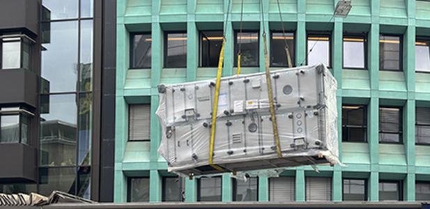 A large air handling unit, wrapped in plastic, is being hoisted up in front of a modern office building using lifting slings