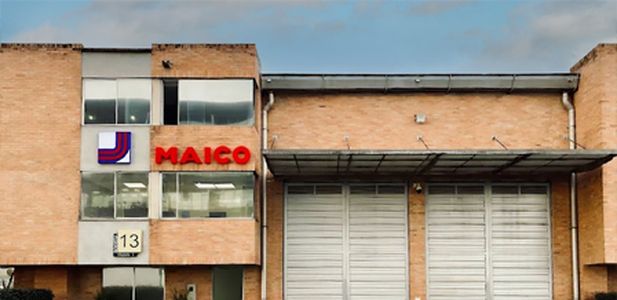 Fassade eines Backsteingebäudes mit großen Fenstern, zwei geschlossenen Rolltoren und rotem Schriftzug 'MAICO' neben einem Logo mit blauen und roten Linien.
