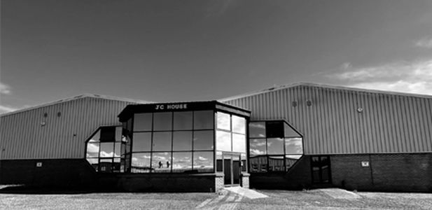 Großes Industriegebäude mit dreieckigem Glasvorbau und Schriftzug JC House und reflektierenden Fenstern unter bewölktem Himmel.
