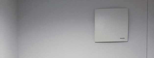 A white, square wall-mounted fan with the word ‘MAICO’ in the bottom right-hand corner, mounted on a light-coloured wall.