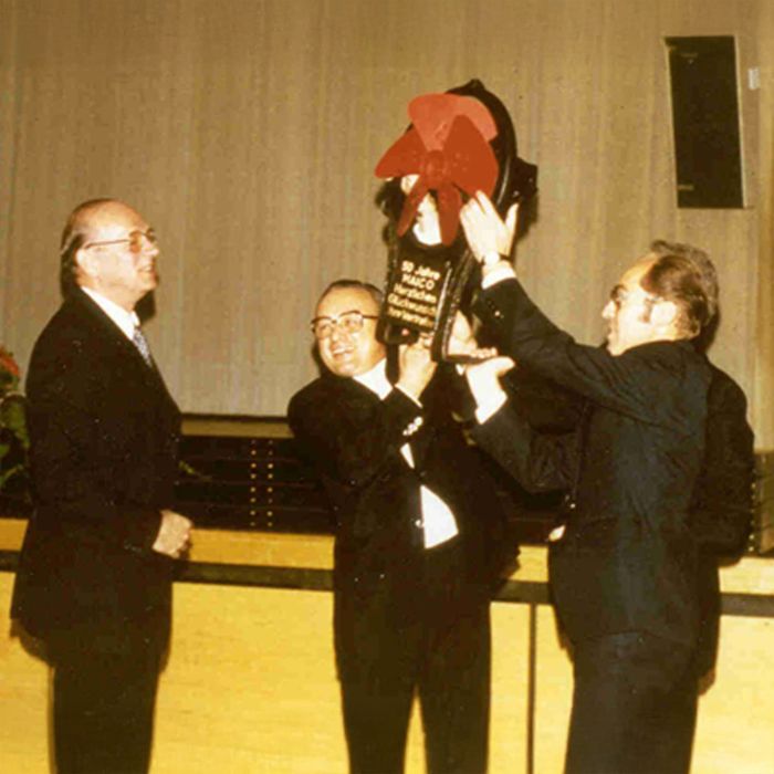 Zwei Männer in schwarzen Anzügen heben einen Ventilator in die Luft mit Aufschrift 50 Jahre MAICO
