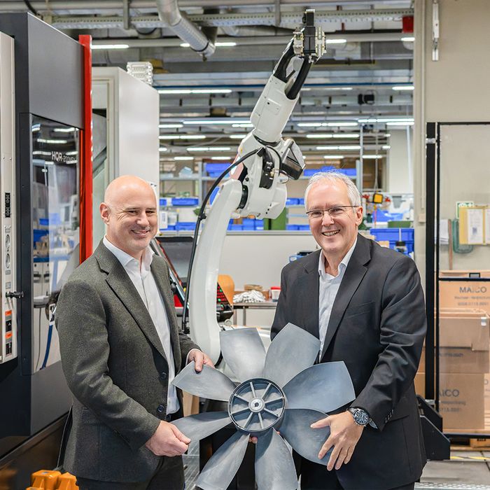 Zwei Männer in Anzügen halten gemeinsam ein großes Flügelrad in einer Fabrikumgebung mit Maschinen und Regalen.