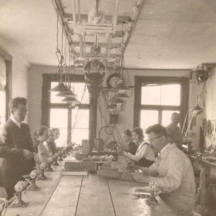 Schwarz-weiße historische Aufnahme von mehreren Personen, die an einem langen Tisch in einem Raum mit großen Fenstern arbeiten.