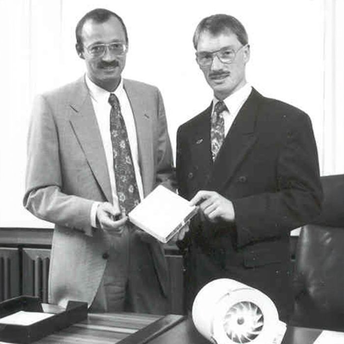 Zwei Männer in Anzügen stehen nebeneinander, einer hält einen Ventilator in der Hand