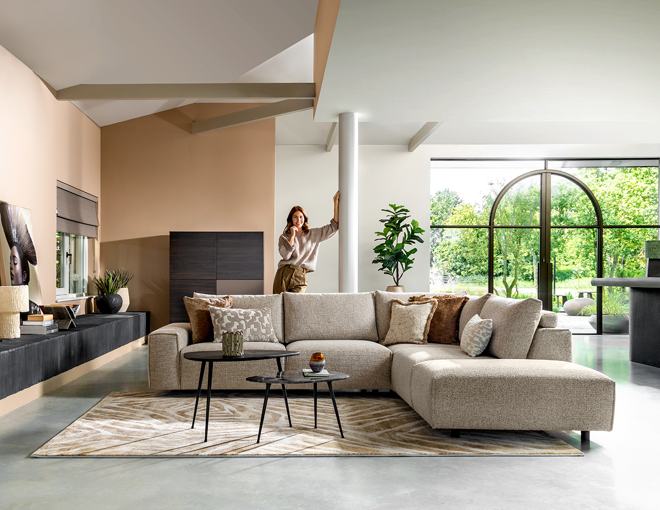 Moderne woonkamer met een beige hoekbank, salontafels, een groot boograam met uitzicht op een groene tuin en een vrouw naast een zuil.
